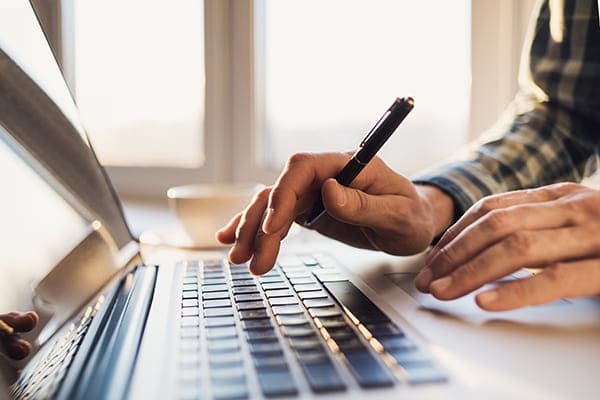 person holding pen and typing on a laptop