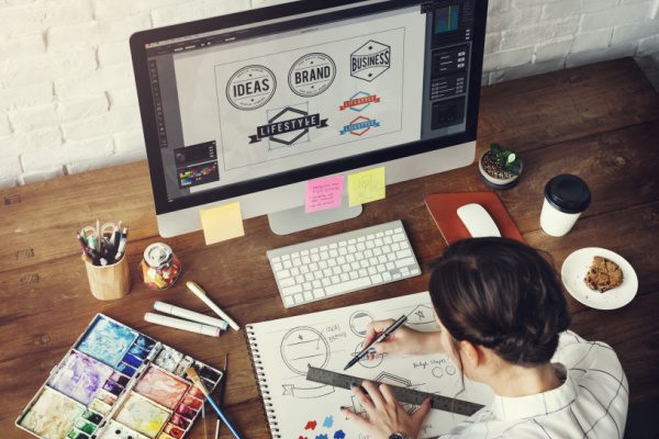 female doing grahic design at a laptop with a notebook and other art supplies to create logos